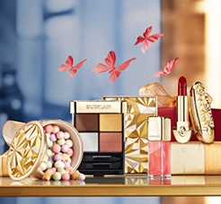 Guerlain sminkkollekció arany csomagolásban: szemhéjpúder paletta, rúzs, szájfény és gyöngyös púder elegáns háttérrel.